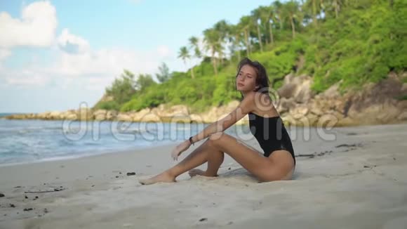 在白色沙滩上与丛林隔离的黑色泳装中身材苗条的黑发模特的肖像视频的预览图