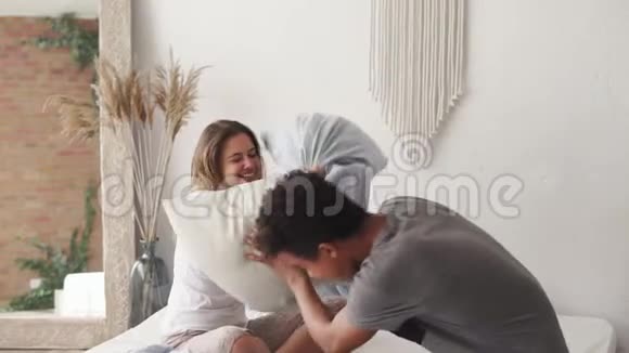 多种族夫妇在床上抱着枕头打架在一起玩得很开心穿着睡衣的女人和男人笑着享受视频的预览图