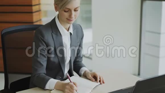 漂亮的金发女商人坐在桌子旁写着笔记本微笑着走进现代办公室的镜头视频的预览图