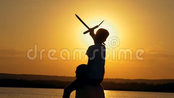 一个幸福的家庭父亲肩上男孩正在玩飞机玩具日落时在湖边剪影视频的预览图