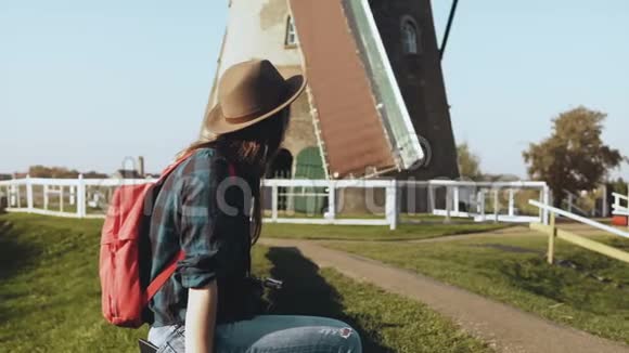 迷人的快乐旅游妇女靠近老风厂美丽的旅行者女孩戴着相机乡村磨坊4K视频的预览图