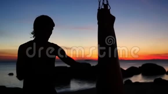 年轻运动员的剪影是拳击在日落时在海滩上用拳击袋练习慢动作视频的预览图