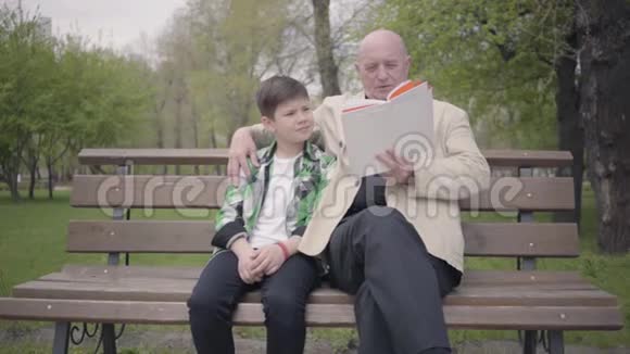 爷爷和孙子坐在公园的长凳上老人为男孩看书世代观念视频的预览图