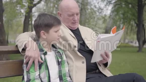 肖像可爱的祖父和孙子坐在公园的长凳上老人为男孩读这本书视频的预览图