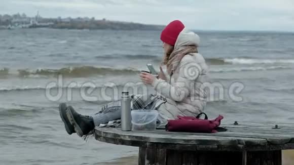 年轻女子穿着暖和的衣服坐在海边坐在木盘管上用热水瓶喝热茶视频的预览图