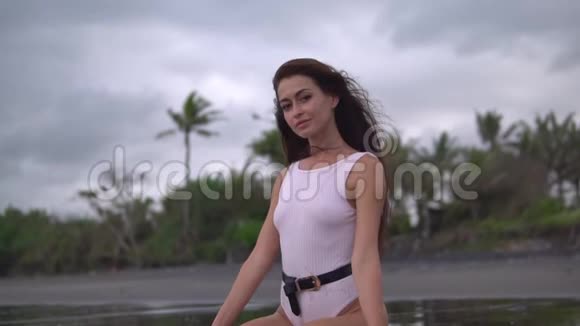 年轻漂亮的女性一头深色卷发身穿白色泳装坐在海边黑色沙滩上视频的预览图