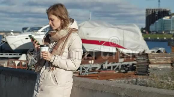 年轻女子穿着白色夹克在河港喝咖啡用电话视频的预览图