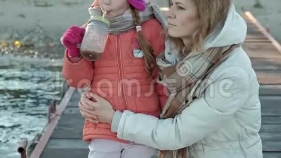 一个女人带着一个女孩一个母亲带着一个女儿坐在河港一个女孩喝着可可热拥抱微笑视频的预览图