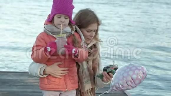 一个女人带着一个女孩一个母亲带着一个女儿坐在河港一个女孩喝着可可热拥抱微笑视频的预览图