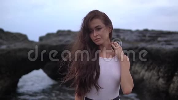 年轻漂亮的女性一头深色卷发身穿白色泳装站在海边的黑色岩石旁边视频的预览图