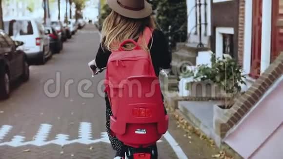 成年女性骑自行车在阳光明媚的城市街道探索兴奋的欧洲女孩骑自行车戴帽子后视图慢速运动视频的预览图