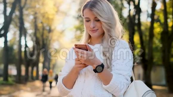 穿白色毛衣的女孩在户外智能手机上写信息女人用数码小玩意在社交网站上滚动视频的预览图