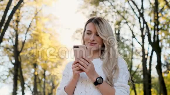 穿白色毛衣的女孩在户外智能手机上写信息的肖像女人用数码小玩意卷轴视频的预览图