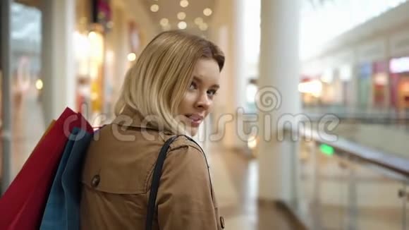 快乐的金发女郎微笑着看着她的肩膀手里拿着购物袋在商场里走来走去视频的预览图