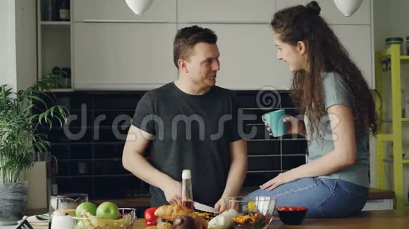 快乐的年轻夫妇在家里的厨房里做饭有魅力的男人一边割一边喂女朋友视频的预览图