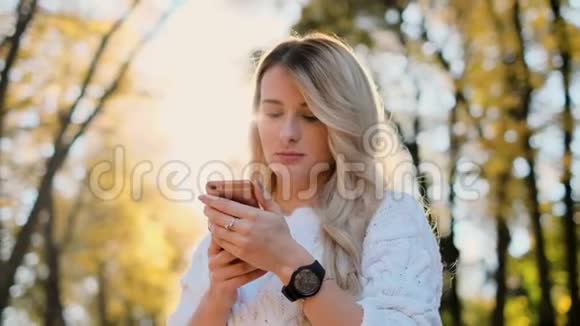穿白色毛衣的女孩在户外智能手机上写信息的肖像女人用数码小玩意卷轴视频的预览图
