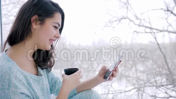 漂亮的女孩坐在互联网上使用智能手机在大窗户附近的家里喝茶视频的预览图