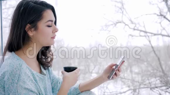 微笑的女性花时间在手机上上网喝茶坐在大的全景窗口附近视频的预览图