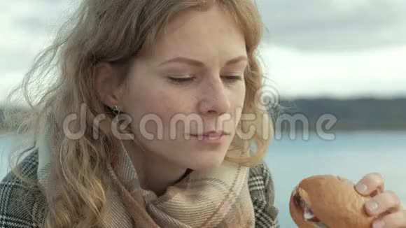 穿着外套的年轻女子坐在河边的海滩上大海野餐在大自然中吃汉堡寒冷的天气视频的预览图