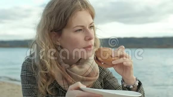 穿着外套的年轻女子坐在河边的海滩上大海野餐在大自然中吃汉堡寒冷的天气视频的预览图
