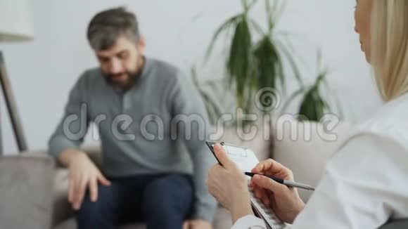 心理治疗师或心理学家在与坐在办公室沙发上的成年男子交谈时写笔记视频的预览图