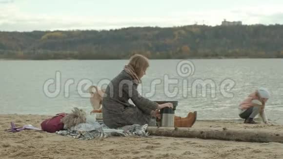 一位穿着外套的年轻女子坐在河边的海滩上大海野餐从热水瓶里倒热茶一杯棕色的视频的预览图