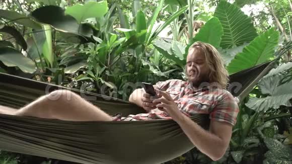 坐在吊床上用智能手机的男人异国情调的背景视频的预览图