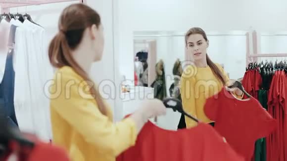 年轻苗条的女士扎着马尾在服装店里挑选一件衣服女人在镜子前购物和检查衣服视频的预览图