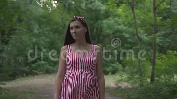 公园里绿树成荫的怀孕女孩一个穿着条纹白红色连衣裙的女孩穿过公园视频的预览图