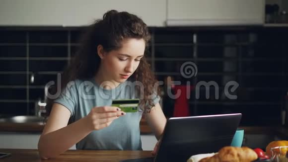 年轻开朗的女性在厨房吃早餐时会使用笔记本电脑和信用卡进行网上购物视频的预览图