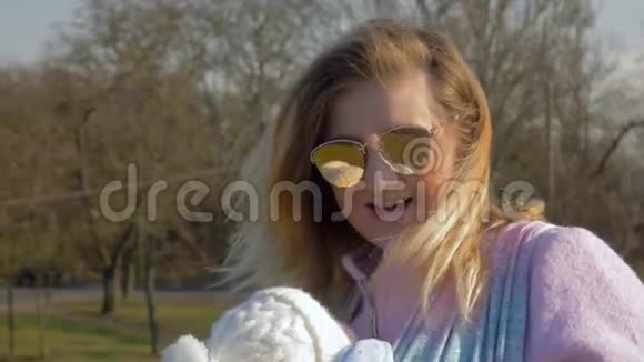 作为母亲年轻的妈妈戴着太阳镜为婴儿唱着歌带着吊带走进公园视频的预览图