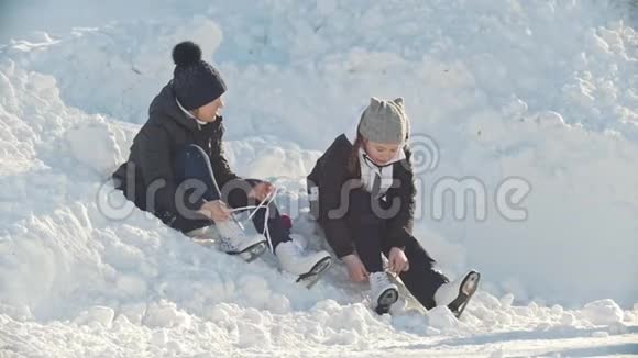 女孩们穿着溜冰鞋坐在靠近溜冰场的雪堆里收紧鞋带动作缓慢视频的预览图
