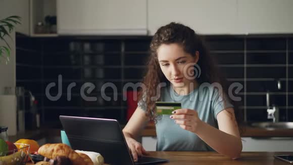 年轻开朗的女性在厨房吃早餐时会使用笔记本电脑和信用卡进行网上购物视频的预览图