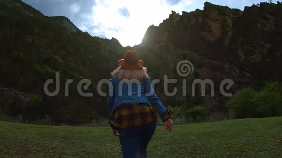 美丽的年轻女子旅行者在大自然中自由地穿越田野在群山把帽子扔到空中视频的预览图