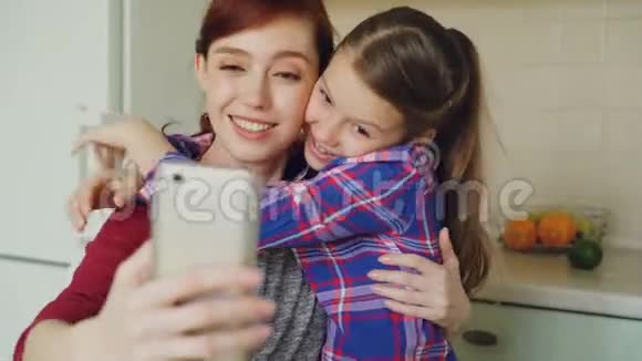 微笑的妈妈和一个有趣的小女儿一起在厨房里用智能手机相机拍摄自拍照片视频的预览图