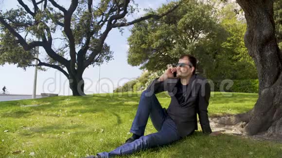 商务人士坐在树旁绿草如茵的草地上在智能手机上聊天视频的预览图