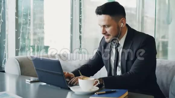 快乐的白人企业家穿着正式的衣服在笔记本电脑上打字而听音乐时耳朵里有耳塞视频的预览图
