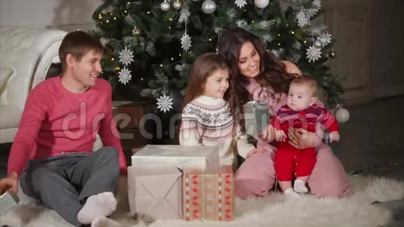 年轻的一家人坐在圣诞树旁的地板上观看新年礼物视频的预览图