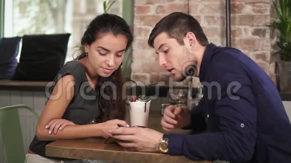 可爱的年轻夫妇牵着手用电话坐在咖啡店的木桌旁浪漫的约会视频的预览图