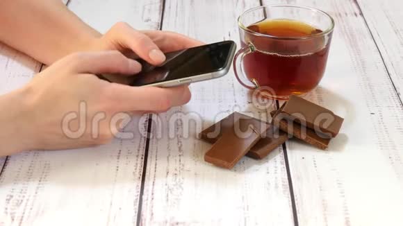一个年轻女人的手拿着智能手机在咖啡馆的特写桌子上有一杯茶巧克力技术视频的预览图