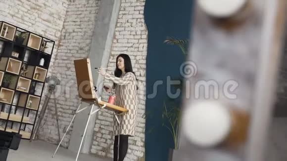 一位穿着连衣裙的年轻女子在画布上画一幅画站在画架后面在一个大气的车间里艺术学院视频的预览图