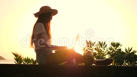 穿着草帽的少女坐在沙滩上在笔记本电脑键盘上打字美丽的热带日落视频的预览图