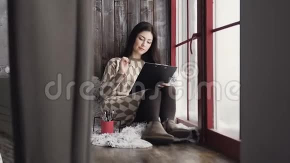 一位年轻的女艺术家坐在窗前的窗台上用化妆品画笔在他的相册里画草图视频的预览图