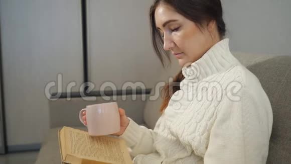 穿着毛衣端着杯茶的年轻女子坐在沙发上看书视频的预览图