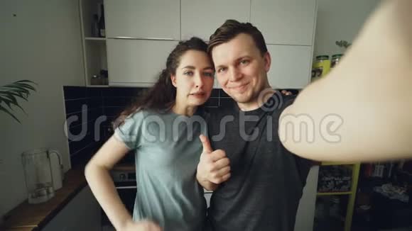 一对年轻有趣的夫妇早上站在家里的厨房里用智能手机相机拍自拍照片视频的预览图