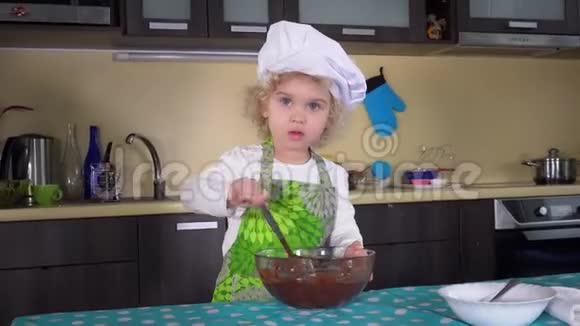 一个严肃的小女孩用勺子把面团混合在厨房的玻璃碗里视频的预览图