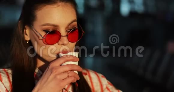 戴着红色太阳镜的迷人年轻女子在傍晚的阳光下喝咖啡视频的预览图