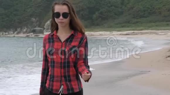 阴天时穿着格子衬衫在沙滩上漫步的年轻漂亮的欧洲女孩视频的预览图