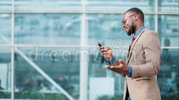 非洲裔美国商人戴着太阳镜在办公楼附近用耳机打电话商务视频的预览图