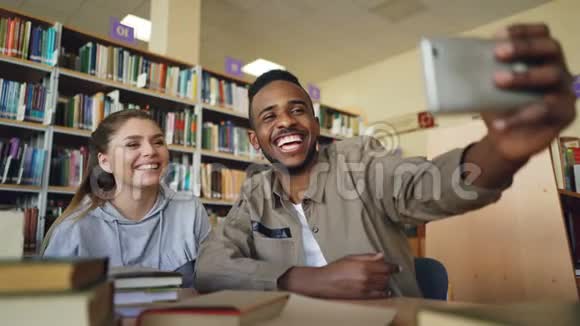 非洲的美国男子和高加索女孩在大学的智能手机摄像头上开心的微笑和自拍视频的预览图